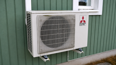 Outdoor unit of a wall-mounted air-to-air heat pump installed beside a house window.
