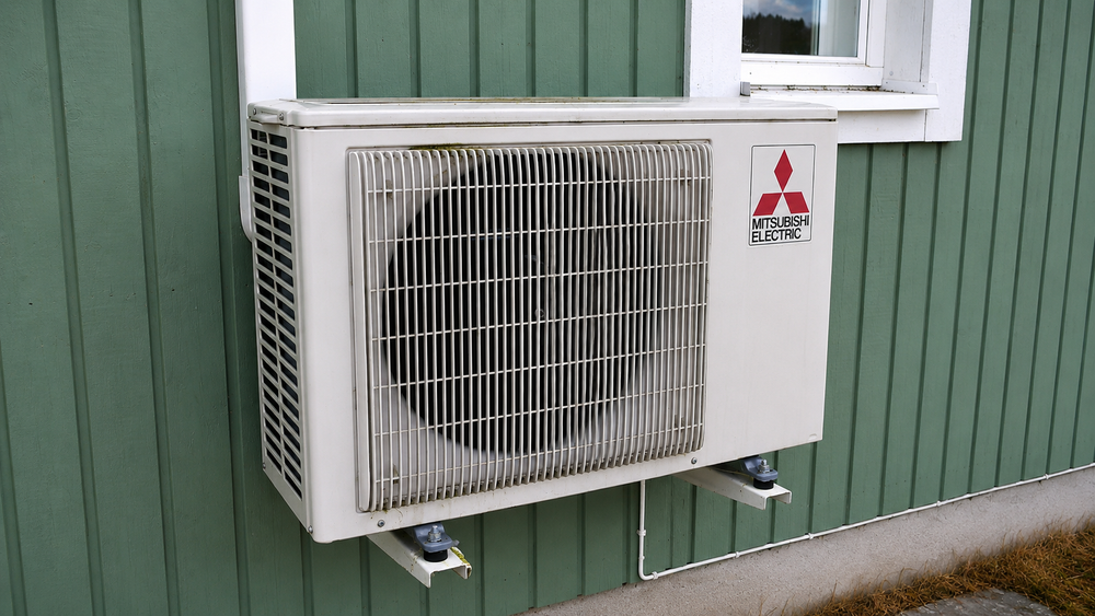 Outdoor unit of a wall-mounted air-to-air heat pump installed beside a house window.